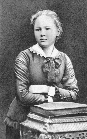 Imagen en blanco y negro de una joven Marie Curie, vista de medio cuerpo, con el cabello recogido y vestida con una blusa formal, apoyada en un pedestal