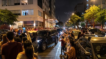 Personas sostienen teléfonos celulares frente a camionetas en una calle nocturna junto a un hotel Marriott y luces en el cielo.