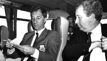 Foto en blanco y negro de Carlos Bilardo, con traje y corbata, leyendo un papel mientras Julio Grondona, también de traje, lo observa en un autobús