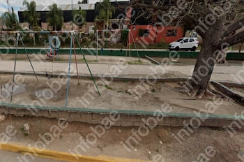 Un parque infantil con columpios, un tobogán, un sendero de cemento, un árbol grande y un vehículo familiar blanco en el fondo
