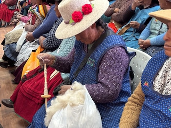 Varias mujeres sentadas; una artesana anciana con sombrero claro y flores rojas, delantal azul y suéter púrpura hila lana con un huso manual