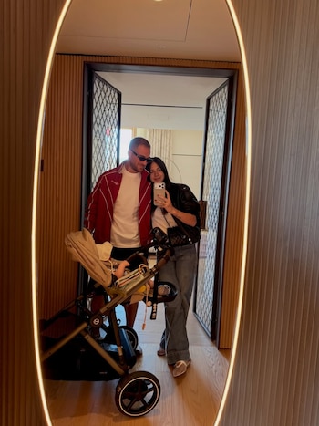 Reflejo en un espejo ovalado de un hombre y una mujer sonriendo, con un bebé en un cochecito al lado. La mujer sostiene un teléfono para tomar la foto