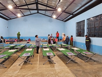Gran salón de albergue con paredes azules y techo de metal, mostrando filas de catres verdes con kits, y varias personas en el fondo