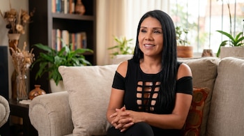 Tula Rodríguez sentada en un sofá claro, vestida de negro, con el cabello largo y oscuro, mirando hacia la derecha con una leve sonrisa. Al fondo, estantes y plantas.