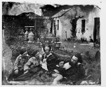 Fotografía en blanco y negro de Alexander Graham Bell con barba, dos mujeres y tres niños sentados en un jardín. Se ve una casa al fondo