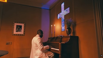 Hombre vestido de traje blanco toca un piano de madera. En la pared, un cuadro con marco dorado, dos velas encendidas, una cruz iluminada y un jarrón con flores