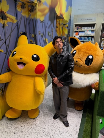 Joe Jonas, con chaqueta de cuero negra, posa entre un Pikachu gigante amarillo a la izquierda y un Eevee gigante marrón a la derecha, en un pasillo con pared floral