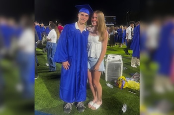 Un joven con toga y birrete azules posa junto a una joven en un campo deportivo durante la noche