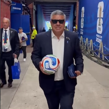 Claudio Tapia, un hombre con traje y gafas de sol, camina por un pasillo sosteniendo un balón de fútbol blanco y azul. Al fondo, se ve un letrero de FIFA World Cup 2026