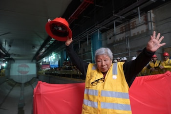 Mujer mayor, chaleco amarillo, levanta casco rojo y una mano. En un túnel de construcción con trabajadores de cascos y una lona roja