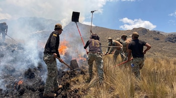 Varias personas con uniformes y palas intentan apagar el fuego y el humo entre la vegetación seca de un cerro.