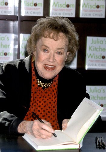 Mujer de edad, pelo rubio, blusa roja y chaqueta negra, sonríe y firma un libro con un bolígrafo negro; libros con el título "Julia's Kitchen Wisdom" al fondo