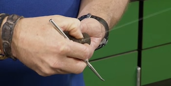 Dos manos masculinas sostienen un sapo de piel rugosa y un instrumento metálico que presiona una glándula, sobre un fondo verde