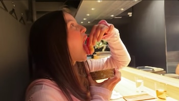 Vista lateral de Emilia Mernes con la boca abierta, recibiendo un trozo de sushi directamente en la boca de una mano, mientras sostiene un cuenco en un bar de sushi