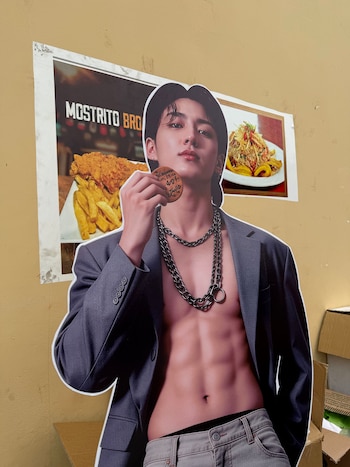 Recorte de cartón de un integrante de BTS posando sin camisa, colocado frente a afiches de comida peruana pegados en una pared amarilla