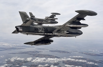 Dos aviones militares de color gris oscuro, con la palabra "ARMADA" en el fuselaje, volando en formación sobre islas y mar bajo un cielo nublado