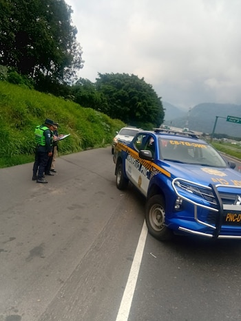 Dos agentes de Tránsito PNC en el borde de una carretera, con una camioneta policial azul y un SUV blanco detrás. Fondo de montañas y vegetación