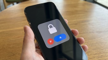 Uma mão segura um iPhone escuro que mostra uma notificação cinza com um cadeado branco, um círculo vermelho com um ‘x’ e um círculo azul com uma seta na tela.