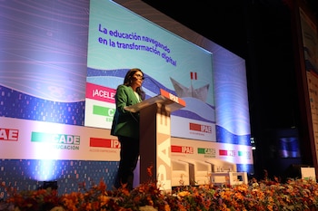 Una mujer con chaqueta verde oscuro habla desde un podio. Detrás, una pantalla exhibe la frase "La educación navegando en la transformación digital" con una ilustración de un barco de papel con la bandera de Perú. El escenario está adornado con flores