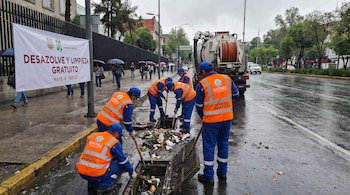 Trabajadores con chalecos naranjas limpian una coladera de una calle mojada. Hay un camión de desazolve y un cartel de "Desazolve y Limpieza Gratuito".