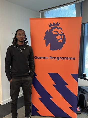 Hombre joven con rastas y vestimenta oscura de pie junto a un banner vertical anaranjado con el logo de un león y el texto 'Games Programme'