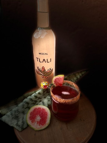 Botella de mezcal Tlali, vaso con cóctel rojo y borde escarchado, rodajas de pomelo, hoja verde y figura de animal sobre madera