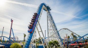Montaña rusa azul Superman: Escape from Krypton con pasajeros en la cima de una torre. Otras atracciones, palmeras y un cielo parcialmente nublado de fondo.
