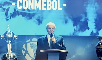 Un hombre de cabello blanco vestido con traje y corbata habla en un podio con un micrófono, frente a una pantalla con el logo de CONMEBOL y dos trofeos