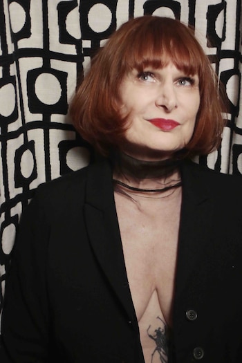 Retrato de una mujer con cabello rojizo, flequillo, labios rojos y un collar translúcido negro, luciendo un blazer oscuro frente a un fondo blanco y negro estampado