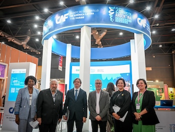 Seis personas, cuatro hombres y dos mujeres, posan sonrientes frente a un gran banner azul circular con el logo de CAF y el texto "25 AÑOS EN ARGENTINA"