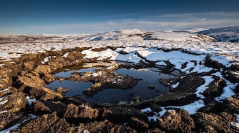 Un terreno hundido con charcos de agua, bloques de tierra oscura y parches de nieve bajo un cielo azul en una llanura.