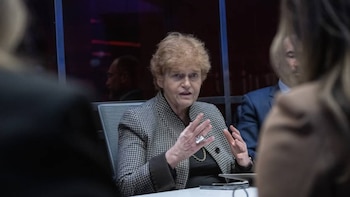 Mujer de cabello rizado y chaqueta a cuadros gesticula al hablar en una reunión; mesa blanca y figuras borrosas en primer plano y fondo oscuro visible