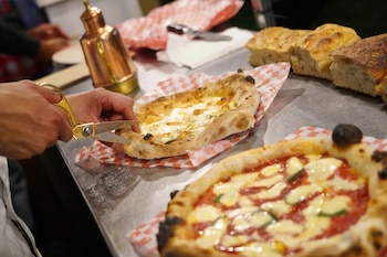 Manos cortan una pizza rectangular de queso blanco con tijeras doradas. Otra pizza redonda, panes y una aceitera de cobre están sobre una mesa metálica