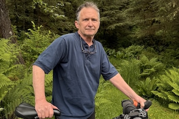 Hombre de cabello gris, con camisa azul y gafas colgadas, de pie junto a una bicicleta. Fondo de bosque con árboles y abundante vegetación verde