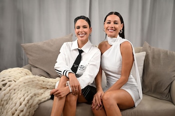 Dos mujeres, Pampita y Barby Franco, se sientan sonrientes en un sofá. Una lleva camisa blanca y corbata negra, la otra un vestido blanco asimétrico. Fondo gris