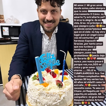 Hombre sonriendo frente a una torta blanca decorada con frutillas y fruta anaranjada, con velas y el letrero 'Feliz Cumple', y un texto conmemorativo a la derecha