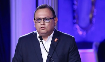 Un hombre de gafas con traje oscuro y camisa blanca habla en un micrófono. Lleva una escarapela roja y blanca en el saco. El fondo es azul borroso