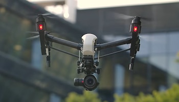 Un dron blanco y negro con cuatro hélices en vuelo, dos luces rojas visibles y una cámara montada debajo. Fondo urbano desenfocado