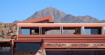 Edificio de líneas horizontales con muros de piedra y cubiertas rojizas, situado frente a una montaña con antenas en la cima