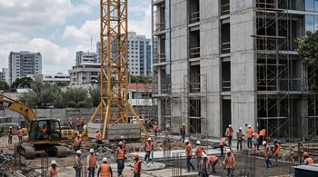 Grúa amarilla y excavadora en obra de construcción urbana. Trabajadores con cascos y chalecos operan junto a un edificio y andamios, con otros edificios y cielo nublado.