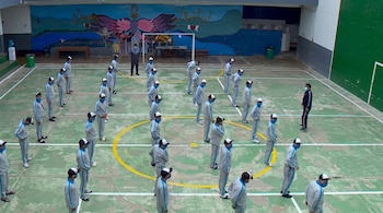 Jóvenes vestidos con buzos grises, gorras y mascarillas azules forman filas en un patio deportivo decorado con un mural al fondo.
