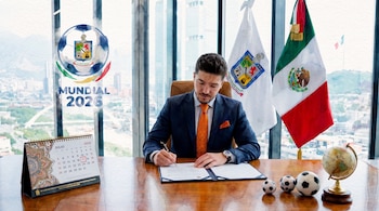 Hombre de traje firma un documento. Banderas de México y Nuevo León, calendario de julio 2026, tres balones de fútbol y un globo terráqueo.