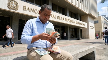 Hombre sentado frente al Banco Central de la República Dominicana contando billetes de pesos dominicanos, con más billetes en su regazo.