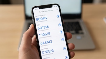 Una mano sostiene un teléfono móvil mostrando la aplicación Google Authenticator con códigos numéricos para Banco Santander, BBVA y CaixaBank.