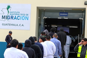 Un grupo de hombres en fila, vistos de espalda, ingresa a un edificio del Instituto Guatemalteco de Migración. Un policía y un guardia están presentes