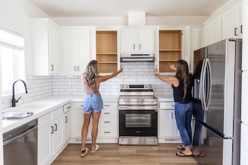 Dos mujeres abren las puertas de los armarios blancos de una cocina equipada con electrodomésticos de acero inoxidable