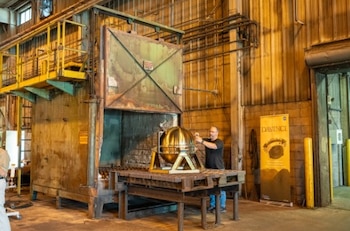 Un hombre ajusta una esfera dorada sobre una plataforma metálica frente a un gran horno industrial abierto y un cartel informativo