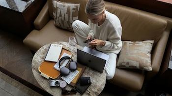 Mujer sentada en sillón de cuero, suéter blanco, usando smartphone. Mesa de centro con laptop Apple, auriculares, libro, cámara, gafas, billetera y vaso.