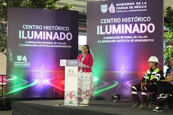 Una mujer habla en un atril ante pantallas que muestran 'Centro Histórico Iluminado' y edificios con luces, mientras un trabajador con casco y chaleco observa