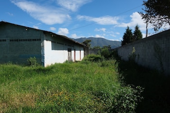 Casa blanca y gris de bloques de cemento en un campo de hierba alta y flores amarillas, con un muro de contención a la derecha y montañas bajo cielo azul